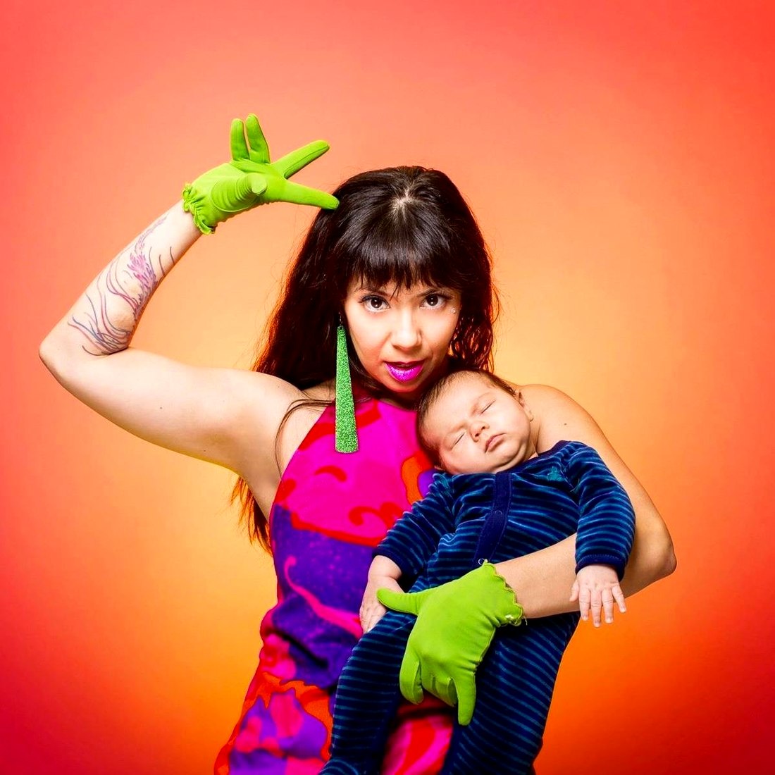 Dork Mom in neon green gloves, flexing one arm in a power pose while cradling a sleeping baby, against a blazing orange backdrop.